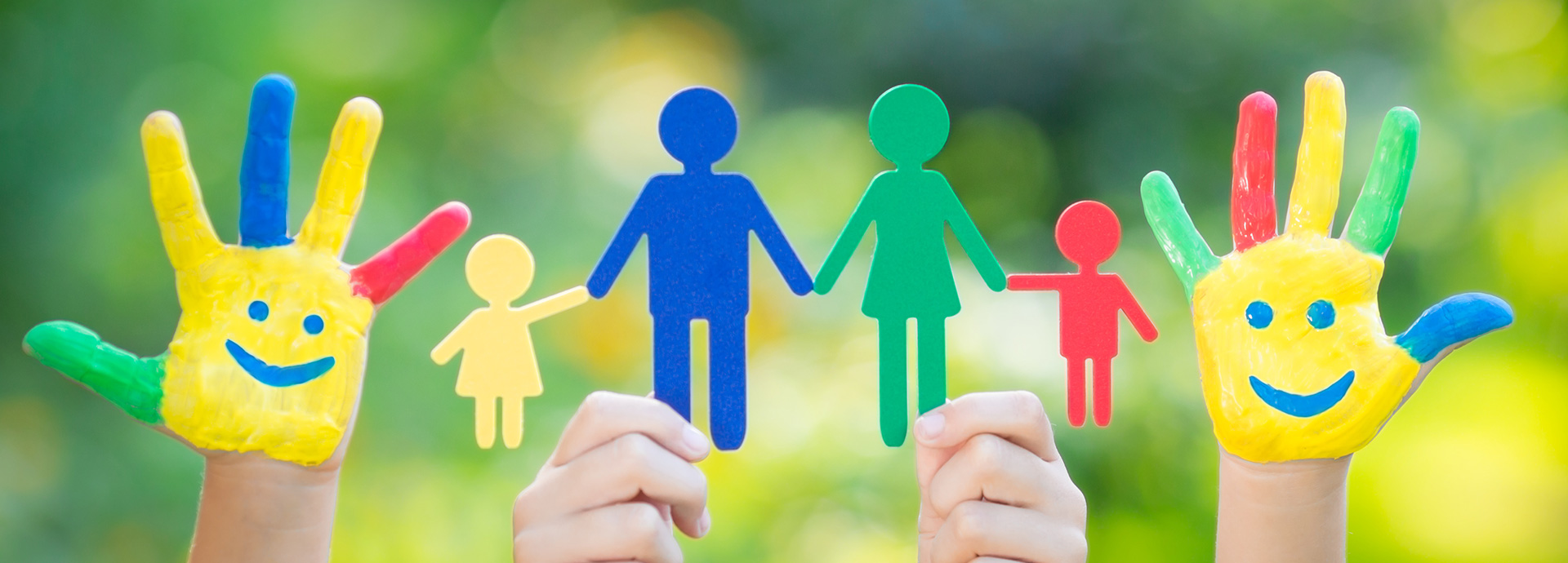 Bunt bemalte Hände halten bunte Figuren einer vierköpfigen Familie im Grünen.