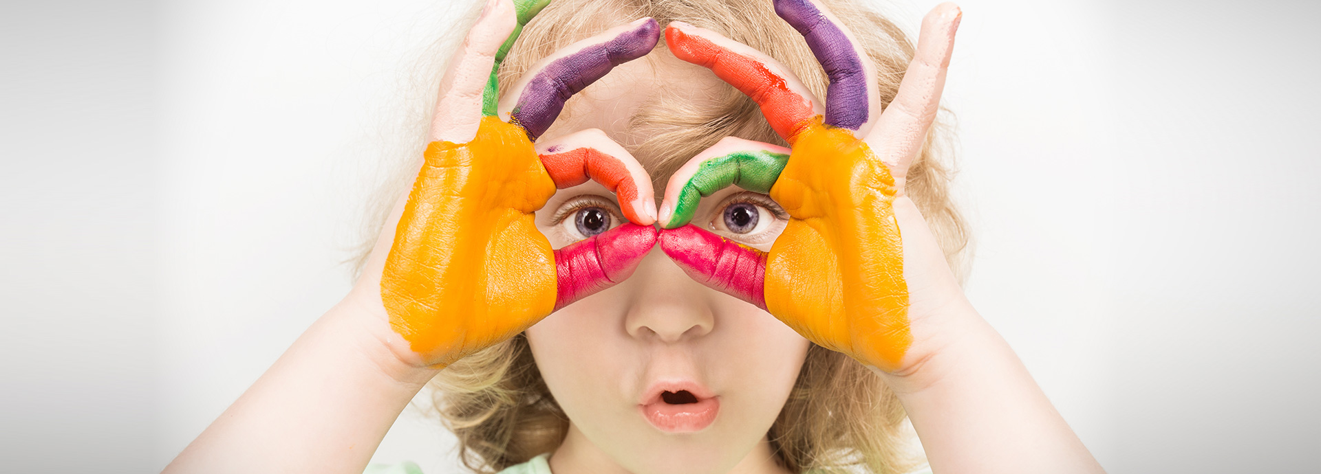 Kind mit bunt bemalten Händen formt eine Brille vor den Augen und schaut überrascht.